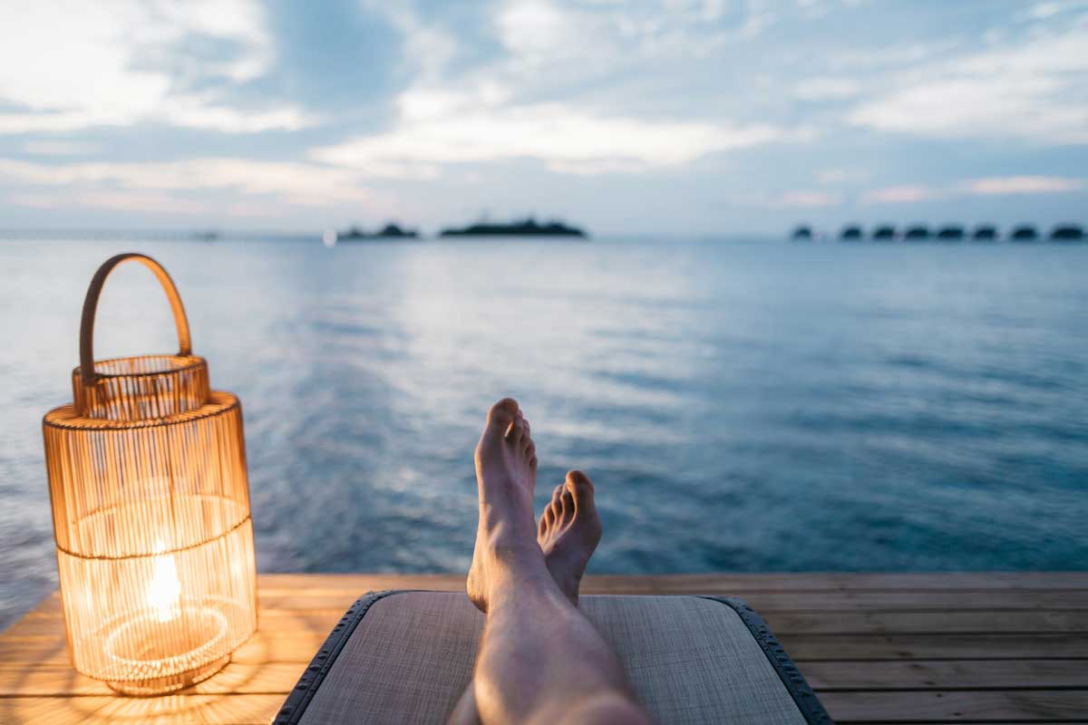 A person's legs are visible on a dock overlooking a wide lake. A lantern sits by their feet, portraying a calm and relaxing environment.